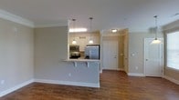 This single-floor space is designed with an open-concept layout that seamlessly connects the kitchen and living areas. The kitchen is a standout feature, equipped with stainless steel appliances, including a French door refrigerator, electric range, and microwave. White cabinetry provides ample storage, complemented by granite-style countertops that extend to a breakfast bar peninsula with seating — ideal for casual dining or entertaining. Two pendant lights hang above the peninsula, adding focused illumination and a decorative touch to the space.

Crown molding runs throughout, lending a polished, finished appearance to the interior. The warm hardwood-style flooring is consistent across the open living and kitchen areas, contributing to a cohesive aesthetic. Neutral gray walls create a versatile backdrop that suits a variety of décor styles.

A hallway visible beyond the kitchen leads to additional areas of the unit, including what appears to be an entryway with a paneled front door. The layout is efficient and well-organized, making good use of the available footprint while maintaining a sense of openness and flow throughout the space.