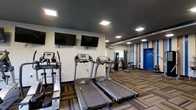 This single-floor fitness center is thoughtfully designed to provide a complete exercise environment. The space features multiple treadmills and an elliptical machine from True Fitness, positioned side by side for convenient cardio workouts. Multiple flat-screen TVs are mounted on the wall above the cardio equipment, allowing users to stay entertained during their sessions. A dedicated strength training area is situated toward the rear of the room, equipped with a multi-functional cable and pulley machine suitable for a variety of resistance exercises. The room is finished with a dark, dramatic ceiling that contrasts with the lighter walls, while recessed square lighting panels provide bright, even illumination throughout. The flooring consists of patterned, multi-tone carpet tiles that add a modern aesthetic to the space. An accent wall featuring vertical stripes in blue, orange, and white adds a vibrant, energetic touch, complementing the bold blue entry door. The overall ambiance is clean, functional, and motivating, making it a practical and visually engaging space for fitness enthusiasts.
