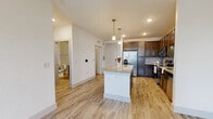 This single-floor unit is designed with a clean, contemporary aesthetic that prioritizes both function and comfort. The open-concept kitchen serves as the centerpiece of the living space, featuring rich dark-toned cabinetry that extends to the upper walls, providing ample storage. A granite-topped island with a white base offers additional workspace and casual seating, while elegant pendant lights hang above, adding a warm, focused glow. Stainless steel appliances—including a French door refrigerator, range, and microwave—complement the white subway tile backsplash for a polished, modern look. Light wood-look plank flooring runs throughout the main living areas, lending warmth and continuity to the open layout. The space transitions into a carpeted bedroom that offers a softer underfoot feel, along with a window fitted with horizontal blinds that invites natural light and outdoor views. The bathroom is thoughtfully appointed with accessibility-friendly grab bars, a granite vanity top, dark cabinetry, and wood-look tile flooring consistent with the overall design palette. Neutral white walls throughout the unit create a bright, airy atmosphere that enhances the sense of openness across every room.
