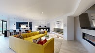 This single-floor amenity space showcases a thoughtfully designed open-concept layout finished with light wood-look tile flooring and crisp white walls throughout. The expansive lounge area serves as the centerpiece, anchored by bold mustard-yellow sectional sofas accented with colorful throw pillows, arranged around a round white coffee table atop a textured area rug. Striped accent chairs add visual variety near the entrance doors, which feature sleek black-framed glass double doors.

A built-in linear fireplace with a wall-mounted television above it creates a striking focal point along one wall, while a tufted olive-green banquette seating nook provides an intimate dining area furnished with a natural wood table, modern white chairs, and dramatic black pendant lighting overhead. Adjacent to this nook is a full kitchenette outfitted with dark cabinetry, stainless steel countertops, a large island with dark stone surfaces, under-counter appliances, and pendant lighting. The kitchen opens directly to an outdoor area through floor-to-ceiling glass doors, inviting natural light deep into the space. Recessed lighting and a wall-mounted split system ensure year-round comfort throughout this stylish communal environment.