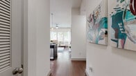 This thoughtfully laid-out one-bedroom, one-bathroom residence offers an efficient and comfortable single-floor plan. A welcoming hallway adorned with wall art leads into the heart of the home, where rich hardwood flooring runs throughout the main living areas. The open-concept kitchen and living room create a seamless, connected space ideal for both everyday living and entertaining. The kitchen features dark cabinetry with granite countertops, stainless steel appliances including a dishwasher, a built-in microwave, and pendant lighting that adds a modern touch. A breakfast bar provides additional seating and blurs the boundary between kitchen and living space. The living room is bright and airy, benefiting from large windows that invite ample natural light and offer views of surrounding greenery. The bedroom is a calm retreat, carpeted for comfort, with dual windows dressed in sheer curtains and access to a dedicated closet complete with shelving. The full bathroom is well-appointed with large-format tile flooring, a dark espresso vanity with granite countertop, wall-mounted cabinetry, and a full tub-shower combination accented with a decorative mosaic tile border. Every detail of this residence reflects a clean, contemporary aesthetic throughout.