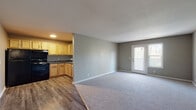 This single-floor unit offers a practical and well-organized layout designed for comfortable everyday living. The open-concept main area seamlessly connects the kitchen and living space, creating a welcoming, airy atmosphere. The kitchen is equipped with warm-toned wood cabinetry, dark laminate countertops, a double-basin stainless steel sink, and black appliances, all set atop durable hardwood-style flooring. The adjacent living area transitions to soft carpeting and is bathed in natural light from a set of glass-panel doors that open to an outdoor space, offering pleasant views of the surrounding greenery. A dedicated entry door provides a clear separation between the main living area and the rest of the unit. The two bedrooms are carpeted and finished in neutral tones, each featuring windows that frame outdoor tree views, lending a calm, natural ambiance. One bedroom includes a built-in closet with a rod and shelving for convenient storage. The bathroom is clean and functional, featuring a full-size soaking tub with a bright white surround enclosure. Throughout the unit, consistent gray-toned walls and white trim provide a cohesive, neutral aesthetic that serves as a versatile backdrop for a variety of interior styles.