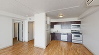 This 1-bedroom, 2-bathroom residence is thoughtfully laid out across a single floor, offering a functional and comfortable living environment. The open-concept kitchen and living area forms the heart of the space, featuring warm hardwood-style flooring that extends throughout the main areas. The kitchen is well-equipped with rich dark-wood cabinetry, light countertops, a stainless steel refrigerator, electric range, built-in microwave, dishwasher, and a stainless steel sink — providing ample storage and workspace for everyday cooking. A structural column subtly defines the transition between the kitchen and the open living area, which benefits from natural light streaming in through large windows dressed with vertical blinds. The bedroom is a comfortable, carpeted retreat with its own window, a wall-mounted HVAC unit, and access to a dedicated closet featuring built-in shelving and a full-width hanging rod for organized storage. Both bathrooms are cleanly finished, with one showcasing classic white penny-tile flooring, a dark-wood vanity, white countertop, and a lighted mirror. A hallway connects the various rooms, contributing to a cohesive and practical flow throughout the residence.