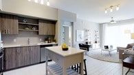 This single-floor unit showcases a thoughtfully designed open-concept layout that seamlessly connects the kitchen and living areas, creating a bright and cohesive living environment. The kitchen is outfitted with two-tone cabinetry featuring dark wood-grain lower and upper cabinets paired with a classic subway tile backsplash. A freestanding white kitchen island with lower shelving and bar seating serves as the central focal point, offering additional prep space and casual dining. Track lighting overhead provides functional illumination throughout the kitchen zone. Stainless steel appliances, including a dishwasher, complement the modern aesthetic. The open living area flows naturally from the kitchen, featuring light-toned tile flooring throughout that enhances the sense of spaciousness. Large windows along the far wall flood the space with natural light and are dressed with sheer curtains. A ceiling fan is centrally positioned in the living area. A lean ladder shelf and framed wall art contribute to the clean, contemporary styling. A doorway visible from the main area leads to an additional room, suggesting a well-connected and functional floor plan overall.