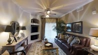 This single-floor space exudes warmth and classic elegance through its thoughtful design and carefully curated details. The living area serves as the heart of the space, anchored by a built-in wall niche that houses both a flat-screen television and a fireplace below, framed by arched shelving that adds an architectural touch. Crown molding runs along the ceiling, lending a refined, polished look to the room.

A ceiling fan with a light fixture sits centrally overhead, providing both comfort and ambient lighting. The vaulted ceiling treatment adds a sense of openness and dimension to the space. Warm, earthy tones on the walls complement the rich hardwood-style flooring, creating a cohesive and inviting palette throughout.

Natural light filters in generously through a large sliding glass door dressed with vertical blinds and elegant drape panels, offering access to an outdoor area. Decorative framed artwork lines the walls, enhancing the space's character. The layout accommodates comfortable seating and a rustic wooden coffee table atop a patterned area rug, tying the room together. Overall, the space balances classic architectural details with a warm, livable ambiance.