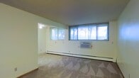 This single-floor space offers an open, functional layout characterized by clean white walls and neutral-toned carpeted flooring. The main living area is generously proportioned, featuring a bank of large windows fitted with horizontal blinds that allow ample natural light to fill the room while providing views of the surrounding trees outdoors. A window-mounted air conditioning unit is positioned along the exterior wall, complementing the baseboard heating system that runs the length of the room — providing year-round climate control options. The space connects to an adjoining area, visible through a wide open passage, which also benefits from natural light via its own window. A ceiling fan is installed in this secondary area, adding additional air circulation. The textured ceiling and straightforward architectural design contribute to a no-frills, practical aesthetic throughout. The layout offers flexibility for various furniture arrangements, and the neutral finishes provide a blank canvas that can be easily personalized. Overall, the space presents a functional and well-lit environment suitable for comfortable everyday living.