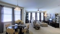 This single-floor residence showcases an open-plan design that seamlessly connects the dining and living areas into one cohesive, welcoming space. The living area is anchored by plush wall-to-wall carpeting in a warm neutral tone, complemented by a comfortable gray sectional sofa, an upholstered ottoman, and a minimalist coffee table. A ceiling fan with an integrated light fixture serves as a functional focal point overhead, while a tall floor lamp adds layered ambient lighting in the corner.

The dining area features a dark-toned round table paired with woven rattan chairs, adding a natural texture to the space. Above it, a pendant light with a brushed nickel finish provides task-oriented illumination. A decorative floral arrangement in a silver vessel serves as a refined centerpiece.

Multiple windows along the exterior wall are dressed with floor-length charcoal gray curtains over white blinds, allowing for excellent light control while contributing to an elegant, cohesive aesthetic. Decorative potted topiaries positioned near the windows introduce a touch of organic greenery. The overall ambiance is calm, polished, and thoughtfully curated.