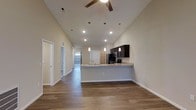 This single-floor residence showcases a thoughtfully designed open-concept layout that maximizes both space and functionality. The living area immediately impresses with dramatically vaulted ceilings that create an expansive, airy atmosphere, complemented by a ceiling fan and recessed lighting for year-round comfort. Warm hardwood-style flooring runs throughout, lending a cohesive and inviting feel to the entire home. The kitchen is a standout feature, boasting rich dark cabinetry, light stone countertops, and a peninsula-style breakfast bar with pendant lighting suspended above — an ideal setup for casual dining and entertaining. Stainless steel appliances, including a refrigerator and range, complete the kitchen's modern aesthetic. A hallway leads from the main living area toward the private quarters of the home, with additional rooms and natural light visible in the distance through interior windows. Neutral-toned walls provide a versatile backdrop, allowing the architectural details — particularly the soaring ceilings and open sightlines — to take center stage. The overall ambiance is clean, modern, and welcoming, making this a highly functional and visually appealing single-level home.