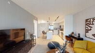 This single-floor residence showcases a thoughtfully designed open-concept layout that seamlessly connects the kitchen, dining, and living areas. Light wood-tone flooring runs throughout, complementing a neutral and contemporary color palette accented by bold blue-gray walls in the living space.

The kitchen is well-appointed with light gray flat-front cabinetry, a subway tile backsplash, stainless steel appliances, and a central island with a white quartz countertop that doubles as a casual dining bar. Track lighting and pendant fixtures illuminate the space effectively. The open dining area flows directly from the kitchen, creating an ideal setting for everyday living and entertaining.

The living room is anchored by a sliding glass door that opens to an outdoor view of lush greenery, allowing generous natural light to fill the room. A dedicated media wall with grey accent paint adds visual contrast and depth.

The bedrooms are well-proportioned and bright, each featuring windows with outdoor views. One bedroom includes a walk-in closet, providing ample storage. The bathroom offers a clean, modern aesthetic with a floating wood-grain vanity, white quartz countertop, large wall mirror, and a tiled walk-in shower.