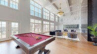 Located at 616 Amelia St in the Overlook neighborhood of Fredericksburg, Virginia, this single-floor amenity space is designed to impress with its grand architectural scale and refined interior finishes. The centerpiece of the space is its dramatically high coffered ceiling, adorned with a tiered gold chandelier that anchors the room with a sense of elegance. Expansive floor-to-ceiling gridded windows line the walls, flooding the interior with abundant natural light and offering views of the surrounding Overlook community.

The open layout is divided into distinct functional zones that flow seamlessly together. A billiards area features a full-size pool table with a red felt surface, positioned beneath the soaring walls accented with wainscoting and soft, nature-inspired mural artwork. The lounge area provides stylish seating, including a large sectional sofa and velvet accent chairs paired with gold-accented side tables, all arranged atop a herringbone hardwood floor.

A sleek bar area is highlighted by floor-to-ceiling dark tile cladding, providing a dramatic contrast to the otherwise bright, neutral palette. Additional tile-clad accent walls add texture and visual depth throughout. An exterior door provides access to an outdoor area, enhancing the overall connectivity of the space.