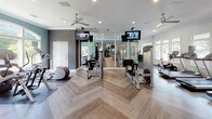 This fitness center is a thoughtfully designed, single-floor facility offering a comprehensive workout environment. The space is defined by its striking herringbone-patterned hardwood-style flooring, which runs throughout and lends a polished, upscale aesthetic. Walls are painted in a sophisticated combination of deep charcoal gray and muted teal, complemented by crisp white trim and crown molding that elevate the overall design.

The main exercise area is generously proportioned and equipped with a variety of strength-training machines, cable systems, and weight benches, alongside functional fitness accessories such as a medicine ball rack. Ceiling-mounted televisions are positioned throughout for convenient viewing during workouts, and ceiling fans provide additional air circulation alongside recessed lighting.

A dedicated cycling studio is separated by a sleek floor-to-ceiling glass partition wall with a metal grid frame, housing a row of professional-grade stationary bikes. Large windows throughout the facility invite abundant natural light and offer views of the surrounding greenery, creating an open and energizing atmosphere. A direct exterior door provides convenient access, and the space is well-maintained with modern safety features throughout.