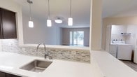 This single-floor, two-bedroom, two-bathroom residence at Premier Indigo Falls offers a thoughtfully designed open-concept layout that seamlessly connects the kitchen and living areas. The kitchen is a standout feature, showcasing crisp white countertops, a mosaic tile backsplash with neutral tones, and a stainless steel sink paired with a modern arc faucet. Pendant lighting above the kitchen counter provides both functional illumination and a contemporary aesthetic touch. A pass-through opening connects the kitchen directly to the living area, enhancing the sense of openness and allowing natural light to flow freely throughout the space. The living area is bright and inviting, highlighted by a sliding glass door that leads to a private balcony, bringing in generous natural light and outdoor views. Neutral wall tones and light flooring contribute to a clean, modern ambiance throughout the residence. The overall design balances practicality with style, making this residence at Premier Indigo Falls a comfortable and visually appealing home.