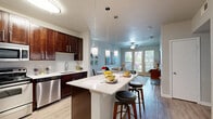 Located in Dallas, TX, this well-appointed one-bedroom, one-bathroom residence offers a thoughtfully designed single-floor layout that maximizes both function and style. The open-concept kitchen and living area serves as the heart of the home, featuring rich dark-wood cabinetry, a subway tile backsplash, stainless steel appliances — including a refrigerator, range, microwave, and dishwasher — and a white quartz peninsula with bar seating, illuminated by modern pendant lighting. The adjoining living room is bright and inviting, with large windows and a glass-panel door that welcome generous natural light and outdoor views, complemented by a ceiling fan overhead and light wood-style flooring that runs throughout the entire unit. The spacious bedroom offers a large window with tree-lined views and direct access to a well-sized walk-in closet. The full bathroom features a soaking tub with shower, dark cabinetry with a quartz-topped vanity, and direct access to the closet for added convenience. A dedicated hallway connects the spaces efficiently. The consistent use of neutral tones, quality finishes, and cohesive design elements creates a clean, modern ambiance throughout.