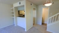 This two-story residence offers a functional and straightforward layout designed for comfortable everyday living. The main living area is finished with wall-to-wall carpeting in a neutral tone, creating a soft and cohesive base throughout the space. A defining architectural feature is the partial wall with a pass-through window that connects the kitchen to the living area, allowing for interaction between the two spaces while maintaining a degree of separation. The kitchen is equipped with cabinetry in a warm wood finish, a dark countertop surface, and standard appliances, all visible through the pass-through opening. Built-in shelving is positioned adjacent to the pass-through, offering convenient display or storage space. The walls are painted in a soft, warm off-white tone that contributes to a clean and neutral ambiance throughout. Ceiling-mounted HVAC vents are integrated throughout the main level, ensuring climate control across the space. Doorways visible from the main living area lead to additional rooms, suggesting a well-connected floor plan. The two-story configuration provides additional living or sleeping quarters on the upper level.