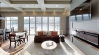 This thoughtfully designed single-floor amenity space offers a seamless open-concept layout that integrates a fully equipped kitchen, dining area, and a comfortable living room into one cohesive environment. The living area is anchored by a plush sectional sofa, a round marble coffee table, and a striking brick-accented fireplace with a wall-mounted TV, creating a warm and inviting focal point. Floor-to-ceiling sliding glass doors flood the space with natural light and provide expansive views of the surrounding outdoor courtyard. A coffered ceiling with recessed lighting adds architectural depth and elegance throughout.

The kitchen is well-appointed with stainless steel appliances, including a large French-door refrigerator and a professional-grade range. A central island with granite countertops and bar seating serves as both a functional workspace and a casual dining spot, illuminated by stylish industrial pendant lights. A textured accent wall behind the kitchen adds a modern design detail. Warm hardwood flooring flows throughout the entire space, contributing to a cohesive and polished aesthetic.

The dining area accommodates multiple seating configurations, ideal for both casual and structured gatherings. Adjacent to the main space, glass-walled private rooms with sliding doors offer dedicated spaces for focused work or meetings, maintaining visual connectivity while providing separation. Overall, this space balances functionality with refined design.
