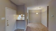 This single-floor residence offers a practical and well-organized layout designed for comfortable everyday living. The interior is finished in warm, neutral tones that create a welcoming ambiance throughout the space.

The kitchen is partially enclosed, featuring a pass-through counter with a granite-style surface that opens toward the dining area, allowing for easy interaction between spaces. It is equipped with white appliances, including a refrigerator, microwave, and range, complemented by wooden cabinetry and a sink positioned beneath a hanging light fixture. Tile flooring grounds the kitchen area with a clean, durable finish.

Adjacent to the kitchen, the dining area is defined by a pendant light fixture that hangs overhead, providing focused illumination. The room transitions seamlessly into the broader living space, which is finished with plush wall-to-wall carpeting that adds warmth underfoot.

The textured ceilings and clean white baseboards throughout the space contribute to a cohesive and tidy aesthetic. Natural light supplements the interior lighting, enhancing the overall brightness of the open layout. The single-floor design ensures easy navigation and accessibility across all areas of the home, making this a functional and inviting living environment.