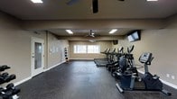 This fitness center offers a comprehensive workout environment thoughtfully designed to accommodate a variety of exercise needs. The space features durable dark rubber flooring throughout, providing both safety and comfort during exercise. A row of True-brand cardio machines — including treadmills and ellipticals — lines one side of the room, each equipped with individual display consoles to track workout metrics. Wall-mounted televisions are positioned for easy viewing during cardio sessions. The open floor area provides ample room for stretching, bodyweight exercises, or functional training. A medicine ball rack along the wall offers a selection of weighted balls for strength and conditioning work. Natural light filters in through a set of windows along the back wall, contributing to an energizing atmosphere. Ceiling fans are installed overhead to promote air circulation and comfort during workouts. Recessed and flush-mounted lighting ensures the space remains well-lit at all times. The neutral-toned walls and clean layout create a focused, distraction-free environment. An entry door with a glass panel provides convenient access, completing this practical and fully functional fitness facility.