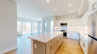 This single-floor unit showcases a thoughtfully designed open-concept layout, where the kitchen and living areas flow seamlessly together. The kitchen is a standout feature, equipped with white shaker-style cabinetry extending to the ceiling, a classic tile backsplash, stainless steel appliances including a range, microwave, and dishwasher, and a generously sized central island with a quartz countertop offering additional workspace and storage. Light hardwood flooring runs throughout the kitchen and living areas, lending warmth to the bright, contemporary palette.

The living area benefits from expansive floor-to-ceiling windows that invite abundant natural light and frame scenic outdoor views of the surrounding neighborhood and green spaces. Bedrooms are finished with plush carpeting, large windows with outdoor views, and direct access to bathroom facilities. The primary bedroom connects to an en-suite bathroom featuring a glass-enclosed walk-in shower, dark-toned vanity cabinetry, and neutral tile flooring. A secondary bathroom mirrors the modern aesthetic with a soaking tub, floating vanity, and clean tile floors. Throughout, the space maintains a cohesive, contemporary design defined by neutral tones, recessed lighting, and quality finishes.