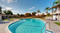 Located at 2970 East Ruby Drive in Fullerton, California, this single-level community amenity space offers a well-appointed outdoor environment designed for recreation and relaxation. The centerpiece of the grounds is a generously sized, freeform swimming pool with crystal-clear blue water, accessible via multiple entry points including staircase rails. The pool deck features lounge chairs, providing a comfortable space to enjoy the Southern California sunshine.

Surrounding the pool, the grounds are thoughtfully landscaped with well-maintained green lawns, mature palm trees, and established shade trees that enhance the resort-like atmosphere. A covered pergola structure offers shaded seating with picnic tables, ideal for outdoor gatherings. A dedicated basketball court sits at the far end of the property, complementing the recreational offerings of the space.

The adjacent multi-story building features covered second-floor balconies with decorative iron railings and exterior staircases, overlooking the pool and common areas below. Wide concrete pathways connect all areas of the grounds, ensuring easy navigation throughout. The combination of lush greenery, recreational facilities, and open sky views creates a vibrant and welcoming community outdoor space in the heart of Fullerton.