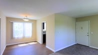 This single-floor space presents a clean, functional layout with a warm and neutral aesthetic throughout. The main living area is finished with smooth hardwood-style flooring that transitions to carpeting in an adjoining zone, offering a subtle distinction between spaces. Walls are painted in soft, warm neutral tones that contribute to a bright and welcoming ambiance.

Natural light enters generously through a large window dressed with white vertical blinds, illuminating the open floor plan and enhancing the airy feel of the interior. A semi-flush ceiling light fixture with a simple, contemporary design provides additional overhead illumination, complementing the natural light effectively.

White baseboards run cleanly along the perimeter of the walls, adding a crisp architectural detail that contrasts neatly with the warm wall tones. An open doorway leads to what appears to be a kitchen or utility area, offering a glimpse of cabinetry and countertop space beyond, suggesting a practical and connected layout. A light switch on one wall indicates additional room connectivity. Overall, the space offers a straightforward, well-maintained interior with a calm and livable atmosphere.