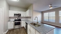 This single-floor residence offers a thoughtfully designed open-concept layout that seamlessly connects the kitchen and living area. The kitchen is a standout feature, equipped with stainless steel appliances including an electric range, over-the-range microwave, and dishwasher. White shaker-style cabinetry with brushed nickel hardware lines the walls, complemented by granite countertops and a classic white subway tile backsplash. A peninsula with an undermount sink and a modern gooseneck faucet provides additional prep space and casual seating potential. Wood-look flooring runs throughout the kitchen and living areas, adding warmth and durability.

The open living area is bright and spacious, benefiting from natural light through a large window with blinds and a ceiling fan overhead. Bedrooms feature plush carpeting, ceiling fans, and generous windows. At least one bedroom includes a sizeable walk-in closet outfitted with wire shelving for organized storage.

The bathrooms mirror the kitchen's design aesthetic, featuring granite-topped white vanities, wood-look flooring, and clean neutral wall tones. One bathroom includes a tub/shower combination with a natural light-admitting window, while another features a single-sink vanity layout. The overall design palette is neutral and modern throughout.