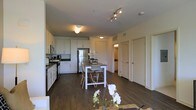 This single-floor residence is designed with an open-concept layout that seamlessly connects the kitchen, dining, and living areas into one cohesive space. The kitchen is outfitted with white shaker-style cabinetry extending to the upper walls, providing generous storage, while stainless steel appliances—including a refrigerator, range, and microwave—complement the modern aesthetic. Dark countertops contrast neatly against the white cabinetry, and the space flows naturally into the dining area, which features a white rectangular table surrounded by light-colored chairs with a woven texture. Dark hardwood-style flooring runs throughout, grounding the interior with warmth and continuity. Track lighting and flush-mount ceiling fixtures illuminate the open areas, while additional rooms visible in the background—accessed through white paneled doors—suggest private living quarters beyond the main living space. The overall ambiance is contemporary and clean, characterized by a neutral color palette of whites and warm wood tones, creating a welcoming and functional environment suited for everyday living.