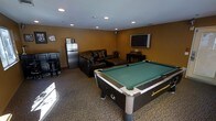 This single-floor entertainment and recreation space is thoughtfully designed for leisure and socializing. The room is finished with warm golden-tan walls and recessed ceiling lighting that creates an inviting, energetic atmosphere throughout. Carpeted flooring adds comfort underfoot while complementing the room's casual, lounge-oriented character.

At the center of the space sits a full-sized pool table with a green felt surface, serving as the focal point for recreational activity. Along one wall, a bar area features a dark wood counter with bar stools, accompanied by framed artwork that adds a personalized touch to the setting. A stainless steel refrigerator and microwave provide convenient refreshment and snack options without leaving the space.

The lounge area is furnished with a plush dark leather sectional sofa accented with decorative pillows, positioned facing a large wall-mounted flat-screen television supported by an entertainment console. This arrangement creates a comfortable viewing and relaxation zone that complements the game area seamlessly.

The combination of the pool table, bar setup, lounge seating, and media display makes this space a versatile and functional entertainment hub, well-suited for gatherings, game nights, and casual downtime. The cohesive layout ensures that all amenities are easily accessible within the room.