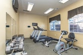 Located at Eastland Court SE in Rome, Georgia, this fitness center offers a well-equipped, single-floor exercise facility designed to support a variety of workout routines. The space is organized to maximize functionality, with distinct areas dedicated to cardio training and strength work. Multiple Spirit-brand treadmills are arranged in a row, complemented by an elliptical trainer and a recumbent stationary bike, providing ample options for cardiovascular exercise. A dedicated free weights area features a multi-tier dumbbell rack stocked with a range of weights, accompanied by a wall-mounted mirror that enhances the sense of space and supports proper form during training. A wall-mounted flat-screen television adds an entertainment element to the workout environment. The room is finished with neutral warm-toned walls and durable carpeted flooring. Natural light filters in through multiple windows dressed with dark valances, offering views of the surrounding outdoor area including what appears to be a pool and landscaping. A glass-panel door provides direct access to the exterior. Overhead fluorescent lighting ensures the space remains well-lit throughout the day. Fitness center rules are posted on the wall, and safety features such as an emergency exit sign are present, reflecting a thoughtfully maintained facility.
