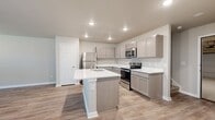 This two-story home offers a well-organized layout spread across two floors. The ground floor houses the garage, which includes an automatic opener, water heater, and direct interior access. The main living level features an open-concept kitchen and living room finished with light wood-tone luxury vinyl plank flooring throughout. The kitchen is outfitted with gray shaker-style cabinetry, white quartz countertops, stainless steel appliances including a refrigerator, electric range, and microwave, and a central island with an undermount sink—providing both additional prep space and casual seating potential. A dedicated laundry room with overhead shelving accommodates a full-size washer and dryer. Upstairs, the home includes three bedrooms, each with closets featuring built-in shelving and hanging rods. The spacious primary bedroom includes a walk-in closet with dual hanging sections. Three full bathrooms are distributed throughout, each featuring gray vanity cabinetry with white countertops and tub-shower combinations. The private backyard is enclosed by a tall cedar wood fence and features a grassy lawn shaded by mature trees, offering a secluded outdoor retreat.