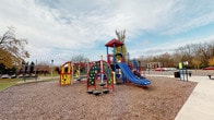 Located at 360 Broad Meadows Blvd in Columbus, Ohio, this outdoor playground offers a well-equipped recreational space designed to engage children of varying ages and abilities. The centerpiece of the area is a large, multi-level play structure finished in bold primary colors — red, blue, yellow, and green — that stands prominently against an open sky backdrop surrounded by mature trees.

The play structure features a variety of interactive elements, including dual blue slides of differing heights, providing options for younger and older children alike. Arch-shaped climbing walls with molded handholds encourage physical activity and the development of motor skills. Additional features include crawl-through tunnels, activity panels, and elevated platforms accessible via multiple entry points, promoting imaginative and social play.

The play area is surfaced with wood chip mulch, offering a safety-conscious ground cover beneath and around the equipment. The open layout of the space allows for clear sightlines across the entire playground, making supervision straightforward. The surrounding environment includes green open lawn areas and mature deciduous trees, contributing to a welcoming, natural atmosphere. A parking area is visible in the proximity, providing convenient access for visitors to this community-oriented outdoor recreational space.