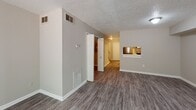 This two-story residence is designed with a functional, neutral aesthetic throughout. The main level features durable wood-look vinyl plank flooring in a cool gray tone, extending through the living area, hallway, kitchen, and bathrooms. The living room is a generously sized open space with a textured ceiling, a sliding glass door that invites natural light and provides exterior access, and a pass-through window connecting to the kitchen. The kitchen is equipped with granite-style countertops, gray cabinetry with brushed nickel hardware, and a stainless steel refrigerator. A half bathroom on the main level includes a vanity with a single sink and wood-look flooring. A carpeted staircase with a white railing leads to the upper floor, where plush carpeting runs throughout the bedrooms. The bedrooms are well-proportioned, featuring textured ceilings, standard closets, and windows that allow natural light. A full bathroom on the upper level includes a vanity with granite-style countertops, a large mirror, and wood-look flooring. The overall color palette consists of warm beige and gray tones, creating a clean and cohesive environment throughout both floors.