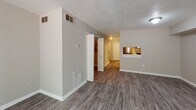 This two-story residence is designed with a functional, neutral aesthetic throughout. The main level features durable wood-look vinyl plank flooring in a cool gray tone, extending through the living area, hallway, kitchen, and bathrooms. The living room is a generously sized open space with a textured ceiling, a sliding glass door that invites natural light and provides exterior access, and a pass-through window connecting to the kitchen. The kitchen is equipped with granite-style countertops, gray cabinetry with brushed nickel hardware, and a stainless steel refrigerator. A half bathroom on the main level includes a vanity with a single sink and wood-look flooring. A carpeted staircase with a white railing leads to the upper floor, where plush carpeting runs throughout the bedrooms. The bedrooms are well-proportioned, featuring textured ceilings, standard closets, and windows that allow natural light. A full bathroom on the upper level includes a vanity with granite-style countertops, a large mirror, and wood-look flooring. The overall color palette consists of warm beige and gray tones, creating a clean and cohesive environment throughout both floors.