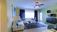 This single-floor space features a well-appointed living room that serves as its central gathering area. The room is finished with light green walls that create a fresh, airy backdrop, complemented by crisp white trim and a smooth white ceiling. A ceiling fan with an integrated light fixture is centrally positioned overhead, providing both air circulation and ambient lighting.

Natural light filters generously into the room through a large picture window framed by bold navy blue curtains, which anchor the space's color palette. A glass-paneled entry door along one wall introduces additional natural light while providing a welcoming point of access. The flooring is carpeted in a neutral tone, and a patterned navy and white area rug defines the seating zone, adding visual interest and texture.

The living area is furnished with a white sectional sofa accented with geometric-patterned throw pillows in navy and white. A sleek rectangular coffee table with chrome legs sits at the center, paired with coordinating side tables featuring glass tops and metallic frames. A large piece of wall art adds a decorative focal point. The overall ambiance is clean, modern, and cohesive.