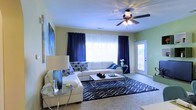 This single-floor space features a well-appointed living room that serves as its central gathering area. The room is finished with light green walls that create a fresh, airy backdrop, complemented by crisp white trim and a smooth white ceiling. A ceiling fan with an integrated light fixture is centrally positioned overhead, providing both air circulation and ambient lighting.

Natural light filters generously into the room through a large picture window framed by bold navy blue curtains, which anchor the space's color palette. A glass-paneled entry door along one wall introduces additional natural light while providing a welcoming point of access. The flooring is carpeted in a neutral tone, and a patterned navy and white area rug defines the seating zone, adding visual interest and texture.

The living area is furnished with a white sectional sofa accented with geometric-patterned throw pillows in navy and white. A sleek rectangular coffee table with chrome legs sits at the center, paired with coordinating side tables featuring glass tops and metallic frames. A large piece of wall art adds a decorative focal point. The overall ambiance is clean, modern, and cohesive.