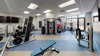 This fitness center offers a comprehensive workout environment designed to accommodate a wide range of exercise needs. The space is organized to maximize functionality, with distinct zones dedicated to cardiovascular training and strength work. Along the windowed perimeter, a row of cardio machines—including treadmills, a StairMaster, and elliptical trainers—face outward, providing users with natural light and views of the surrounding cityscape during their workouts.

The strength training area is equally well-appointed, featuring a multi-station cable machine at the center of the floor, a selection of selectorized strength machines, and adjustable benches for free weight exercises. A rack stocked with medicine balls, kettlebells, and stability balls of varying sizes supports functional and core training routines. A wall-mounted flat-screen television adds an entertainment element to the space.

The flooring consists of a durable rubber and vinyl composite in neutral tones with blue accent sections, offering both safety and visual definition between zones. Recessed panel lighting in the drop ceiling ensures consistent, bright illumination throughout. The overall design is clean and modern, with white walls and a textured accent wall providing a polished, purposeful aesthetic.