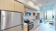 This contemporary residential unit is designed with a clean, modern aesthetic, featuring light wood-tone cabinetry, light hardwood-style flooring, and exposed concrete ceilings that lend an industrial character throughout. The fully equipped kitchen is outfitted with stainless steel appliances — including a refrigerator, electric range, built-in microwave, and dishwasher — complemented by light wood cabinetry extending to the ceiling for ample storage and a sleek countertop with an undermount sink. The shared bedroom spaces are thoughtfully arranged, each accommodating twin beds with built-in drawer storage beneath, individual desks with task chairs, and open closets providing dedicated storage for each occupant. Generous floor-to-ceiling windows flood the sleeping areas with natural light while framing sweeping elevated views of the surrounding neighborhood and tree canopy. The living area offers a comfortable sofa positioned beside a large window that continues the panoramic outlook. The full bathroom is finished with light wood vanity cabinetry, a large mirror, and a full bathtub with shower combination. Consistent use of light tones and natural materials throughout the unit creates a bright, cohesive, and functional living environment well-suited for shared occupancy.