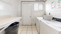 Located at 10237 Western Ave in Downey, California, this single-room utility space serves as a dedicated laundry and mechanical room. The interior is finished with white-painted walls featuring a lower wainscoting detail, and the floor is covered in light-toned ceramic tile throughout. The room is equipped with commercial-grade Speed Queen washers and dryers, providing coin-operated laundry functionality. A large water heater with exposed copper piping occupies one corner, supplying the building's hot water needs. A wall-mounted folding table offers a convenient surface for sorting and folding laundry. Natural light enters through a barred window near the entry door, supplementing the interior lighting. Posted notices and instructional signage are displayed on the walls. A standard door with a knob provides access to the space, which opens to a corridor connecting to the building exterior. The room's compact, efficient layout accommodates all essential utility functions while maintaining clear circulation space. Overall, the space presents a functional, no-frills environment designed to meet the practical laundry and mechanical needs of the building's occupants.