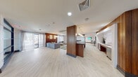 This contemporary amenity lounge is thoughtfully designed to offer a welcoming and functional communal experience. The single-floor space is anchored by light wood-look tile flooring that runs throughout, creating a clean, cohesive foundation that complements the rich, warm-toned vertical wood slat accent walls featured prominently across multiple zones.

The entry foyer greets visitors with frosted glass doors, a pair of soft curved accent chairs, and large black-framed windows that invite natural light. Moving deeper into the space, a dramatic built-in media wall displays a mounted flat-screen TV flanked by a decorative metallic accent display and a pair of deep blue velvet swivel chairs, forming an intimate conversation nook.

The open lounge area features a generously sized sectional sofa in neutral gray, accented with yellow and navy throw pillows, centered around a marble-top coffee table. A cluster of pendant lights hangs overhead, while sheer floor-to-ceiling curtains frame access to an outdoor terrace beyond.

A stylish wet bar with a dark stone countertop, marble surround, and pendant lighting over gold-framed bar stools serves as a social centerpiece. Adjacent to this is a secondary lounge and dining zone with a wall-mounted television and bar-height seating. Textured wallcovering lines a connecting corridor, completing this polished and well-appointed space.