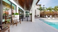Located at 4970 SW 124th Ave in Miramar, Florida, this resort-style pool area delivers a refined outdoor living experience designed for both relaxation and social gathering. The centerpiece is a generously sized swimming pool with crystal-clear water, framed by expansive large-format porcelain tile decking that extends throughout the space. A substantial covered pavilion defines the heart of the area, featuring a striking wood-plank ceiling with recessed lighting that adds warmth and texture to the contemporary design. Floor-to-ceiling glass doors with dark metal frames line the pavilion's interior-facing wall, providing seamless visual connectivity between covered and open-air spaces while flooding the area with natural light. Horizontal-rail metal fencing adds a sleek, modern aesthetic to transitional zones. Comfortable outdoor lounge chairs and seating are thoughtfully arranged beneath the covered area, offering shaded respite from the Florida sun. Textured white stone columns and facades provide architectural contrast against the clean white exterior walls of the surrounding building. Mature palm trees and tropical landscaping create a lush, resort-like ambiance, while decorative planters with manicured topiaries add polished finishing touches. The overall design blends modern architecture with South Florida's outdoor lifestyle seamlessly.