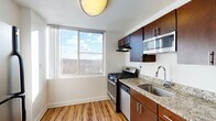 This well-appointed single-floor unit offers a thoughtfully designed layout with a bright, open living area at its core. Rich wood-style flooring extends through the main living and kitchen spaces, complemented by white walls and a textured ceiling that together create a clean, neutral backdrop. Large windows flood the living area with natural light and frame expansive views of a treetop canopy and open sky, indicating an elevated position within the building.

The kitchen is efficiently laid out and features dark espresso cabinetry paired with granite countertops, a stainless steel gas range, dishwasher, microwave, and a double-basin sink. A window positioned above the countertop area continues the theme of natural light and scenic outdoor views.

Additional rooms include a spacious bedroom with plush carpeting and its own large window offering the same elevated outdoor views, as well as a secondary room with wood-style flooring and track lighting. The bathroom is finished with a dark wood vanity, white solid-surface countertop, a large mirror, and tile flooring. The overall ambiance throughout the unit is bright, clean, and modern.