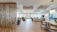 Located at 200 Dominion Oak Circle in Cary, NC, this single-story community clubhouse offers a generously sized, thoughtfully designed gathering space that blends comfort with contemporary style. The open-plan living room serves as the heart of the facility, showcasing soaring coffered ceilings, warm hardwood-style flooring, and an elegant chandelier. A striking white stone fireplace with a wood mantel anchors one end of the room, creating an inviting focal point surrounded by stylish seating arrangements in neutral and coastal blue tones. Large multi-pane windows and French doors flood the space with natural light while offering views of the outdoor amenity area. The kitchen features white cabinetry, granite countertops, stainless steel appliances, and a center island with bar seating beneath pendant lighting. A dedicated game and lounge area is outfitted with a billiards table, a long communal farm-style table with leather bar stools, and patterned blue carpeting that adds texture and warmth. Wainscoting and crown molding throughout lend a refined, classic character to the space. The office provides a quieter retreat, while a slatted wood partition elegantly defines zones within the open layout. Overall, the space exudes a polished yet welcoming coastal-inspired ambiance ideal for community gatherings.