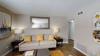 This single-story residence offers a thoughtfully organized layout with distinct living, dining, kitchen, and sleeping areas. The living room serves as the central gathering space, finished with wood-look flooring and neutral gray walls that create a warm, cohesive atmosphere. A large decorative mirror anchors one wall, while a front-facing window allows natural light to fill the room. Accent chairs and a sofa arrangement provide ample seating, complemented by coordinating gold and yellow tones carried throughout the space.

The dining area is situated adjacent to the living room and accommodates a full dining table, with bold artwork adding visual interest to the corner nook. The kitchen is well-equipped, featuring white cabinetry, granite-style countertops, a gray subway tile backsplash, and black appliances including a range, built-in microwave, and dishwasher. Wood-look flooring continues into this area, reinforcing a consistent design flow.

The primary bedroom offers carpeted flooring and neutral walls, providing a calm retreat. A secondary bedroom, also carpeted, includes a window with natural light and closet access. The bathroom features a white vanity with a granite-style countertop, a mirror, and coordinating fixtures, accessible via a hardwood-floored hallway.