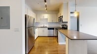 Located at 14307 Greenwood Ave N in Seattle's Greenwood neighborhood, this one-bedroom, two-bathroom residence offers a clean, modern aesthetic defined by bright white walls, polished hardwood flooring, and an efficient single-floor layout. The open-concept living, dining, and kitchen area forms the heart of the space, filled with natural light from large floor-to-ceiling windows. The kitchen is well-appointed with white shaker-style cabinetry, gray quartz-style countertops, a subway tile backsplash, and a full suite of stainless steel appliances including a refrigerator, range, dishwasher, and microwave. A peninsula counter provides additional prep space and a natural divide between the kitchen and dining area. The living room opens directly to a private balcony through a sliding glass door, offering outdoor views of surrounding greenery. The bedroom is comfortably sized and also features a sliding glass door that brings in abundant natural light. Both bathrooms are finished with large-format light tile flooring and clean, minimal fixtures. A dedicated laundry area with side-by-side washer and dryer adds everyday convenience. A hallway with dark-trimmed doors connects the rooms, lending a contemporary contrast to the crisp white walls throughout.