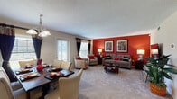This single-floor residence offers a thoughtfully designed layout that balances comfort and functionality across multiple living spaces. The open-plan living and dining area serves as the heart of the home, where a bold red accent wall anchors the living room, complemented by plush carpeting, a wall-mounted flat-screen television, and a round glass-topped coffee table. The adjoining dining area features a dark wood table with upholstered cream chairs, a multi-bulb chandelier overhead, and a door that allows in generous natural light. The kitchen is well-equipped with warm maple cabinetry, dark granite-style countertops, a striking mosaic tile backsplash, tile flooring, and black appliances including a dishwasher and range. The primary bedroom is generously sized with soft carpeting, warm neutral walls, dual windows, and ample space for a king-sized bed flanked by matching nightstands and lamps. A versatile secondary room, currently styled as a den or home office, features carpet flooring, a large window, and classic framed artwork. The full bathroom includes a single vanity with a large framed mirror, a tub-shower combination, and decorative wall accents that tie together the home's warm, coordinated color palette throughout.