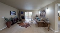 This single-floor residence offers a thoughtfully designed layout with a cohesive neutral color palette of warm beige and taupe tones carried throughout. The living room serves as a central gathering space, featuring plush carpeting, a sliding glass door that opens to an outdoor area, and generous wall space accented by decorative lighting and bold artwork. The open feel of the room is enhanced by natural light from the sliding door.

The kitchen is well-equipped with light wood cabinetry, granite-style countertops, recessed lighting, and white appliances including a range, microwave, and refrigerator. A built-in breakfast bar with seating adds functional dining space, and an adjacent laundry area provides added convenience.

The primary bedroom is generously sized with soft carpeting, a large window, and multiple doorways connecting to closets and additional rooms. Two additional bedrooms offer comfortable accommodations, each with carpeted floors, natural light, and ample space.

Two full bathrooms feature tile flooring, light wood vanities with granite-style countertops, and tub-shower combinations. Textured ceilings with flush-mount lighting are consistent throughout, contributing to a clean, uniform aesthetic across the entire residence.