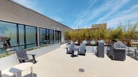 This rooftop terrace offers a well-appointed outdoor retreat with a contemporary design aesthetic. The open-air space features a light-toned concrete deck that provides a clean, modern foundation throughout. Strategically placed large planter boxes are filled with tall bamboo and ornamental shrubs, adding a natural, verdant element that creates a sense of privacy and greenery against the urban backdrop.

Seating areas are thoughtfully arranged across the terrace, including charcoal-gray Adirondack chairs paired with a round side table, as well as additional bench seating — all ideal for relaxation or socializing. A stainless steel BBQ grill positioned near the rooftop's edge makes the space well-suited for outdoor entertaining and gatherings.

Floor-to-ceiling glass doors and large windows along the adjacent building façade connect the interior to the outdoor space, allowing abundant natural light to flow through. The rooftop is enclosed by a sleek metal railing that ensures safety while preserving the expansive views.

The standout feature of this terrace is its unobstructed panoramic view of the Los Angeles skyline, with the downtown high-rises, surrounding neighborhoods, and distant mountain ranges all clearly visible. The open sky above and the cityscape below combine to create a vibrant and inviting rooftop environment.