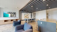 Located at 13639 George Junction in Surrey, BC, this thoughtfully designed amenity lounge offers a refined and versatile gathering space across a single floor. The interior is immediately distinguished by a dramatic slatted ceiling feature finished in deep charcoal tones, which anchors the open-plan layout and adds architectural depth throughout the space.

The lounge is organized into two distinct zones. The first is a relaxed living area furnished with plush grey sofas, upholstered armchairs, and a round coffee table, all set atop a textured area rug. Floor-to-ceiling windows flood this zone with natural light and frame views of the surrounding urban landscape. A wall-mounted large-screen TV paired with a warm oak media credenza completes this comfortable setting.

The second zone transitions seamlessly into a dining and kitchen area. A long communal dining table with modern chairs sits beneath elegant pendant lights, ideal for group gatherings. Adjacent to it, a fully equipped kitchen features sleek grey cabinetry, a stainless steel sink, an induction cooktop, and a generous quartz-topped island with bar seating.

A cozy nook with built-in bench seating, a decorative textile wall panel with bold geometric accents, and open shelving in warm wood tones adds an intimate retreat within the space. Warm hardwood flooring, curated artwork, and recessed lighting throughout contribute to the space's polished, welcoming ambiance.