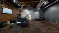 Located at 1110 Locust St in downtown St. Louis, this single-floor amenity space masterfully combines historic character with contemporary design. The open layout is anchored by exposed brick walls featuring elegant arched details, raw wooden ceiling beams, and visible industrial ductwork — a nod to the building's architectural heritage. Soaring ceiling heights enhance the spacious, airy feel throughout.

The main lounge area is furnished with plush blue sofas and green accent chairs arranged around low coffee tables, creating comfortable seating vignettes ideal for socializing or relaxing. A wall-mounted flat-screen TV and a ceiling fan add functional touches to the living area, while an eye-catching chandelier serves as a decorative centerpiece overhead.

A dedicated recreation zone features a full ping-pong table and a oversized Connect Four game, offering casual entertainment options. Patterned carpet tiles in warm earth tones ground the space and add visual texture underfoot. A secondary room provides a dining or meeting area with table-and-chair seating, alongside a beverage station with refrigerators.

A separate flex room with a wide entry door and sidelight windows offers additional versatile space. Track lighting and ceiling-mounted spotlights ensure well-distributed illumination throughout, while large sliding panel partitions allow for flexible spatial configurations.