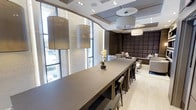 This single-floor space showcases a refined contemporary design, combining functionality with upscale aesthetics. A large, sleek dining or conference table serves as the centerpiece, surrounded by stylish bar-height chairs and accented by a small orchid arrangement, adding a subtle touch of natural elegance. Overhead, a series of cylindrical drum pendant lights hang at varying heights, contributing to the layered lighting scheme alongside recessed ceiling fixtures.

The space benefits from generous natural light streaming through tall, floor-to-ceiling windows framed in dark metal, which also offer views of the exterior environment. A dramatic curved glass wall element adds architectural interest and visually expands the space. The ceiling features tiered coffers with integrated lighting, adding depth and dimension to the overall design.

Toward the rear, a plush lounge area is defined by a deeply upholstered, padded accent wall in dark chocolate tones, paired with neutral sectional seating and decorative cushions. The light-toned flooring contrasts effectively with the darker furnishings, creating a balanced, sophisticated ambiance throughout. The overall design reflects a polished, professional environment with a welcoming atmosphere.