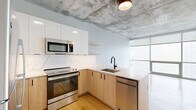 This thoughtfully designed 2-bedroom, 1-bathroom unit spans a single floor and showcases a compelling blend of industrial and contemporary aesthetics. Throughout the space, light wood flooring and exposed concrete ceilings create a striking visual contrast that defines the unit's character.

The open-concept living area seamlessly connects the kitchen, dining, and living spaces. The kitchen is a standout feature, equipped with stainless steel appliances including a range, microwave, refrigerator, and dishwasher. Warm natural wood cabinetry is paired with crisp white upper cabinets, a marble-look quartz backsplash, and matching quartz countertops. Matte black fixtures add a modern, cohesive touch. Floor-to-ceiling windows flood the living area with abundant natural light and offer views of the surrounding exterior.

The bedrooms are well-proportioned, with the primary bedroom featuring its own exposed concrete ceiling detail and access to natural light. Three closets throughout the unit provide ample storage.

The full bathroom is finished with a light wood vanity, white quartz countertop, dual undermount sinks, matte black hardware, a glass-enclosed shower, and neutral tile flooring. A dedicated laundry area and central hallway complete the functional layout of this well-appointed urban unit.